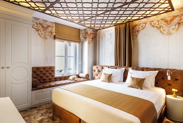 A bedroom with a large bed, a seating area with a cushioned bench, a wardrobe, and a decorative ceiling.