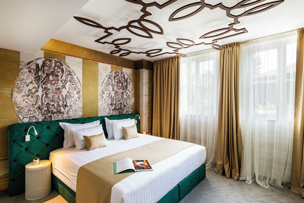 Quarto de hotel com uma cama grande, cabeceira estofada em verde, teto decorativo e cortinas leves.