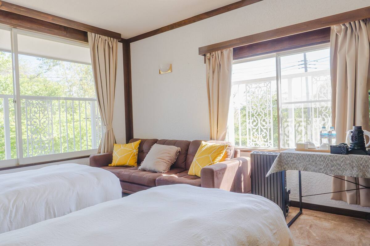 Une chambre d’hôtel avec deux lits, un canapé, une table recouverte d’une nappe et de grandes fenêtres avec des rideaux.