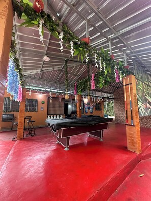 Un espace extérieur abrité comprenant une table de billard, des chaises et des décorations florales suspendues.