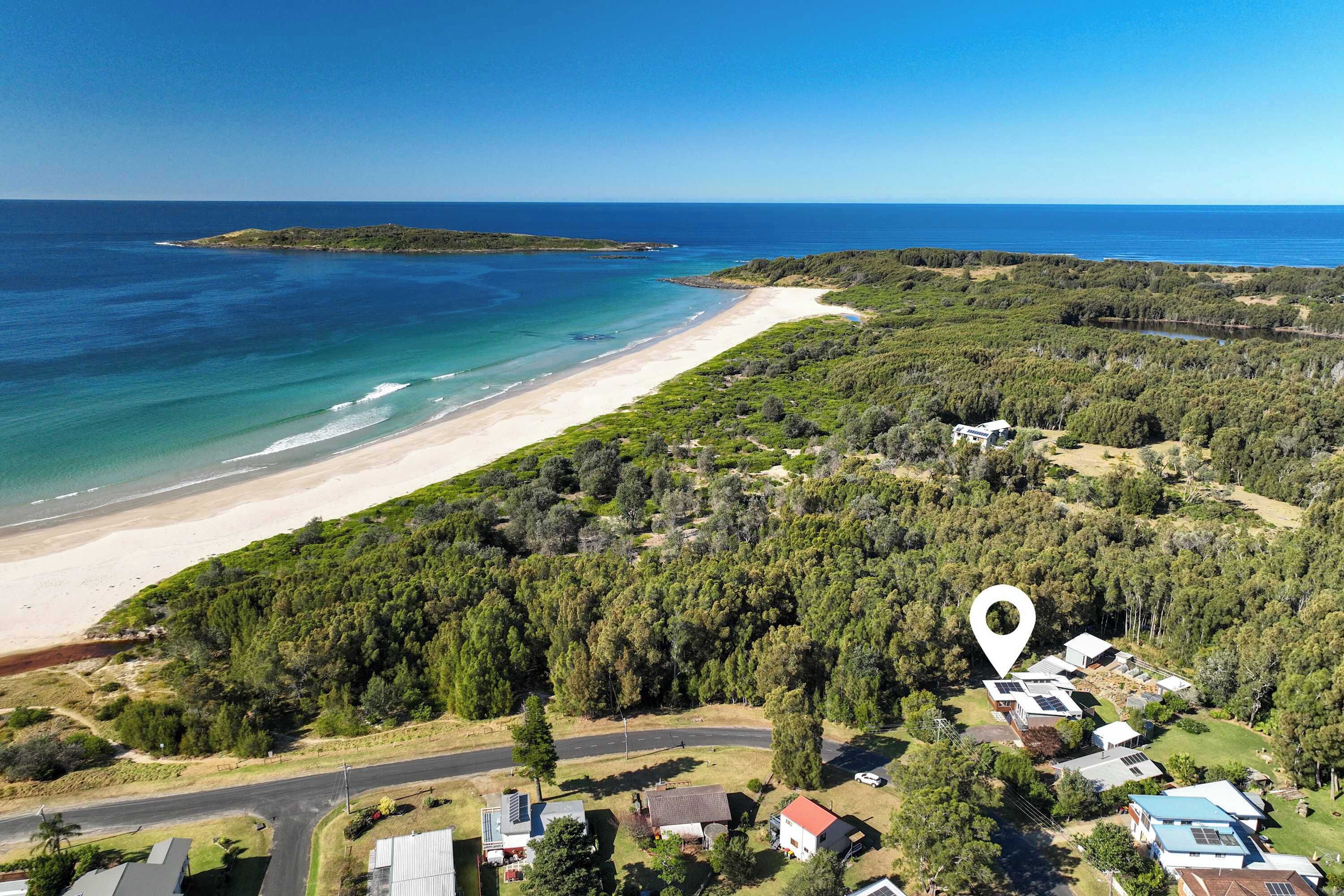 An aerial view of a coastal area with a beach, forested land, and several buildings including a house with solar panels.
