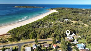 Eine Vogelperspektive zeigt eine Küstenregion mit Strand, bewaldetem Land und mehreren Gebäuden, darunter ein Haus mit Solaranlage.
