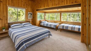 A cabin with a large bed, two smaller beds, and a window with a view of trees. - The Fibro Majestic and Coastal Escape (Bawley Point)