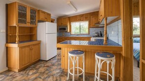 A kitchen with wooden cabinets, a white refrigerator, a microwave, a gas stove, and two stools. - The Fibro Majestic and Coastal Escape (Bawley Point)