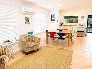 A modern living room with a patterned armchair, a dining table with colorful chairs, a kitchen area with a window, and a rug on the floor. - Narrawallee Holiday House (Narrawallee)