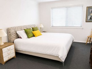 A bedroom with a bed, bedside tables, a lamp, a window with blinds, and a framed picture on the wall. - Narrawallee Holiday House (Narrawallee)