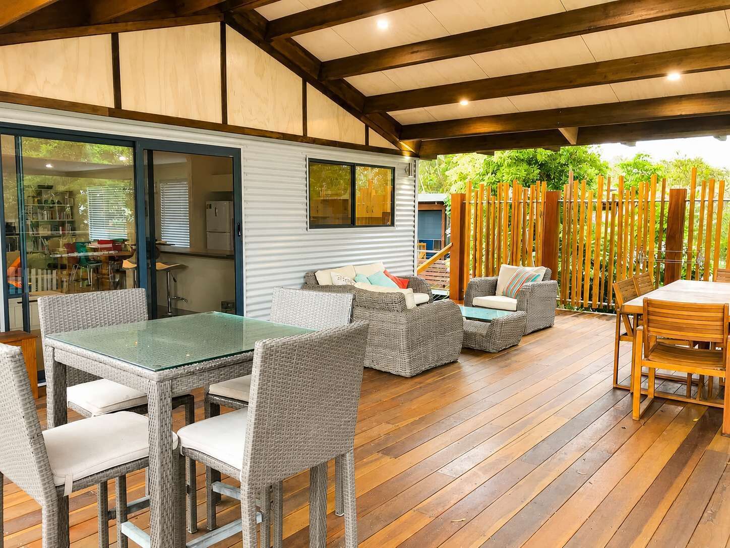 An outdoor patio area with a dining table and chairs, a covered seating area with cushions, and a view of a garden through a glass door.