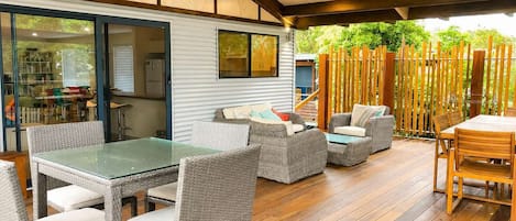 An outdoor patio area with a dining table and chairs, a covered seating area with cushions, and a view of a garden through a glass door.