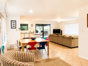 A modern living room with a sofa, a dining table with chairs, a television, and a kitchen area with a window. - Narrawallee Holiday House (Narrawallee)