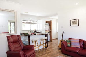 A living room with a red sofa, a red armchair, and a wooden coffee table. There is a kitchen area with a refrigerator, microwave, and a window. - Sandy's Beach Shack at Bawley (Bawley Point)