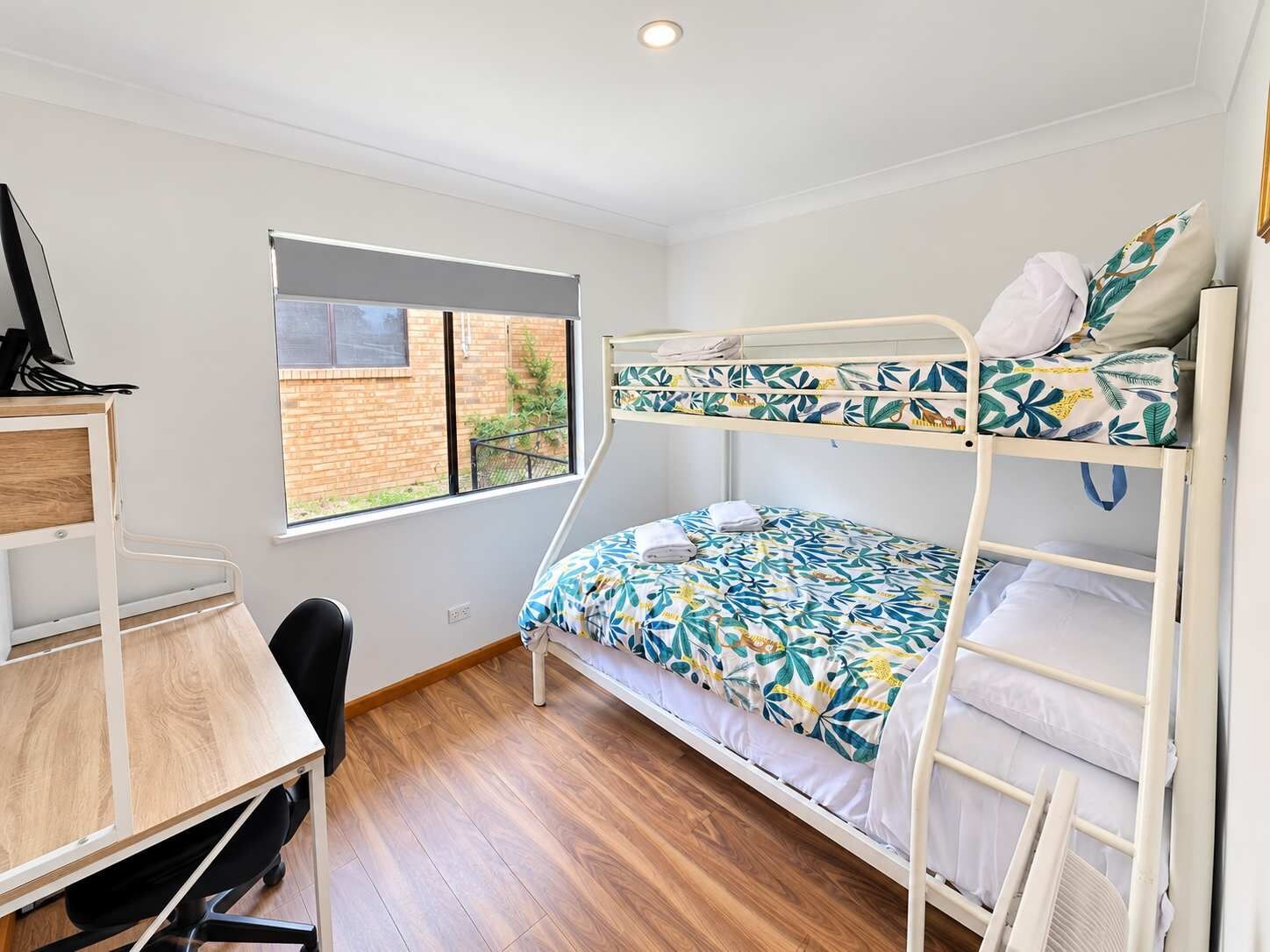 A bunk bed room with a desk and chair, a window with a view of a brick building, and a ceiling fan.