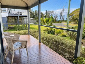 A covered outdoor area with a wooden deck, wicker furniture, and a view of a landscaped garden and road.