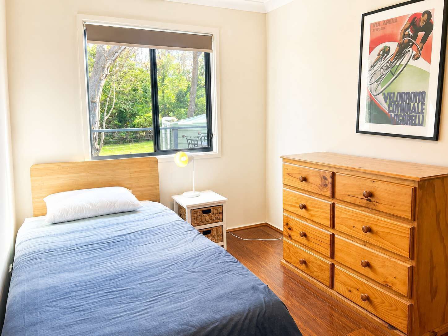 Un dormitorio con cama, cómoda, mesita de noche, lámpara y ventana con vistas a follaje.