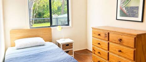 Un dormitorio con cama, cómoda, mesita de noche, lámpara y ventana con vistas a follaje.