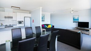 A modern kitchen with white cabinets, a microwave, and a stainless steel range hood. There is a dining area with black chairs and a glass table. A black sofa with colorful cushions is in the living area, and a TV is on a wooden stand. - Fathoms 19 at Mollymook (Mollymook Beach)