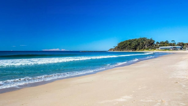 A beach with clear blue waters, a sandy shore, and a building on a small hill in the background. - Fathoms 19 at Mollymook (Mollymook Beach)
