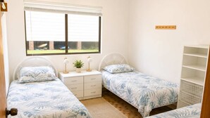 Une chambre avec deux lits simples, une table de chevet avec des lampes, une fenêtre avec des stores et une petite étagère.
