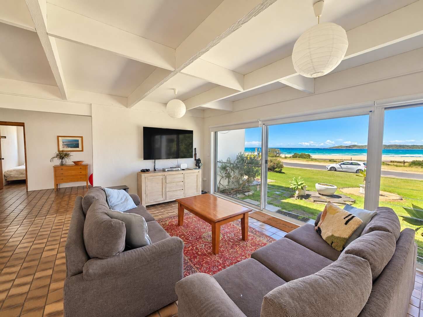 A modern living room with a large window, a flat-screen TV, and a view of the beach.
