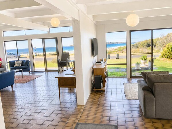 Ein großzügiger Wohnbereich mit einem großen Fenster mit Blick auf einen Strand, ein Essbereich mit Tisch und Stühlen sowie eine Sitzecke mit einer Couch.