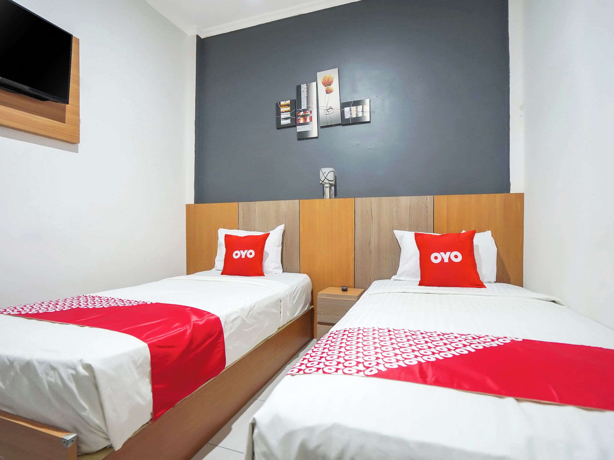 A hotel room with two beds, each with a red and white bedspread, and a grey wall with artwork above.