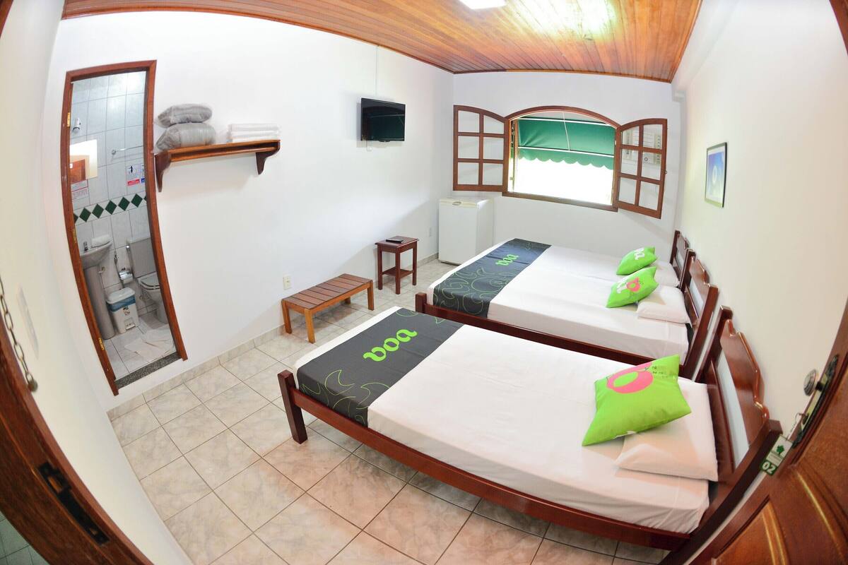 Una habitación con dos camas, una mesita, un espejo y una ventana con una persiana abierta.