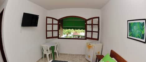 A small room with a bed, a desk, a chair, a TV, and a window with an open shutter.