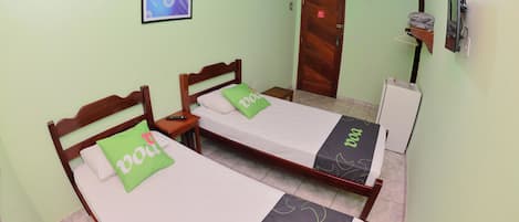 A room with two wooden beds, each with a green and white bedspread, a small bedside table, a wooden door, and a wall-mounted TV.