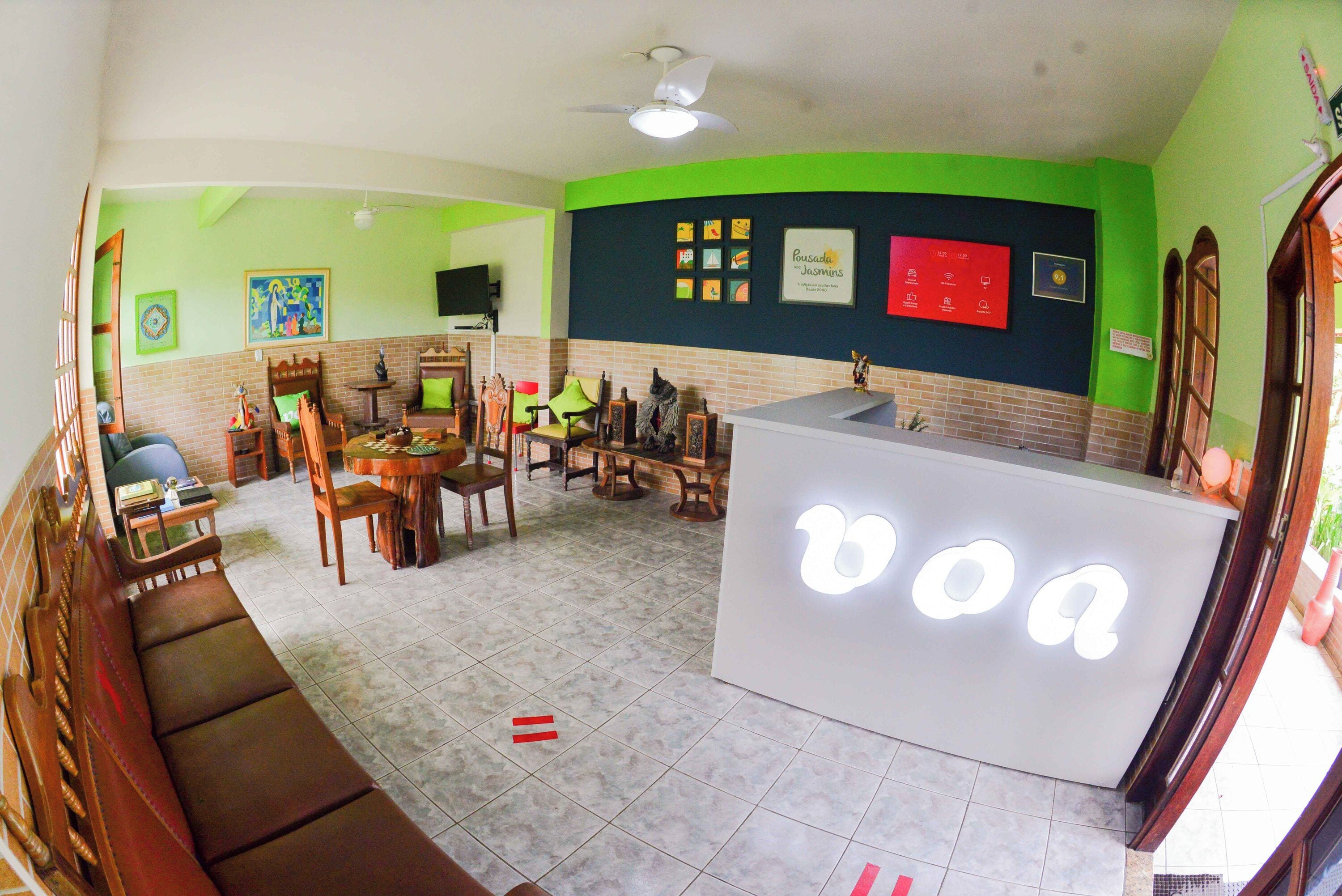 A room with a reception desk, a sofa, a dining table with chairs, and a television on the wall.