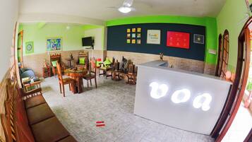 A room with a reception desk, a sofa, a dining table with chairs, and a television on the wall.
