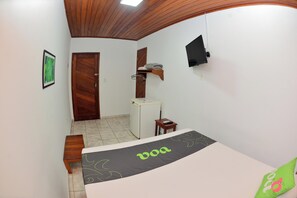 A small room with a bed, a desk, a chair, a TV, a door, and a refrigerator. - VOA Pousada Dos Jasmins (Serra)