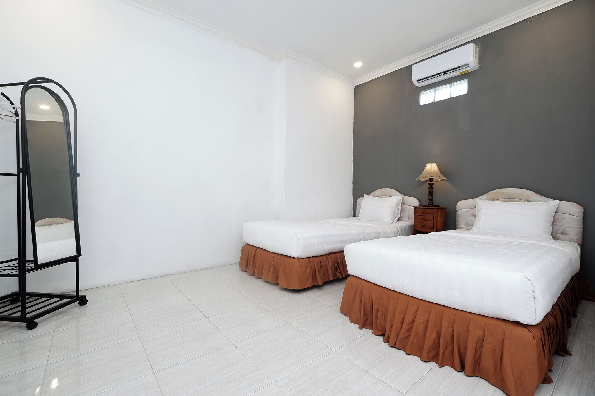 A hotel room with two beds, a standing lamp, an air conditioning unit, and a standing shelf.