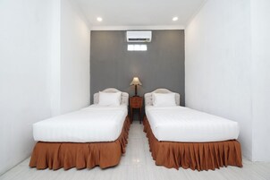 A hotel room with two beds, a small table with a lamp, an air conditioning unit, and a window.