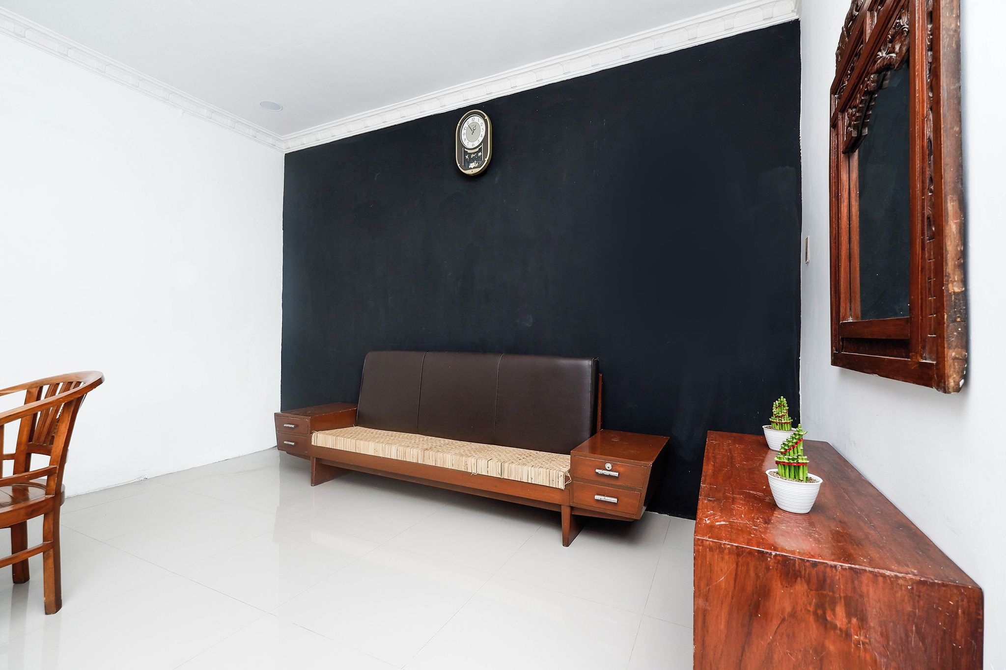 Una habitación con pared oscura, un sofá de madera con cojín, una silla de madera, una mesita con planta en maceta y un espejo en la pared.