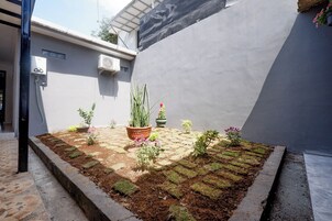 Um pátio com vasos de plantas, um pequeno canteiro e um edifício com parede cinzenta e aparelhos de ar condicionado.