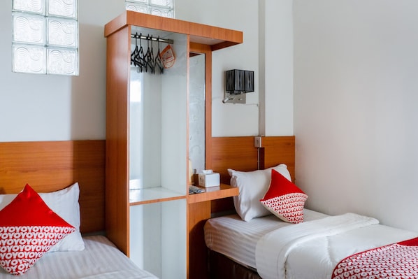 Habitación de hotel con dos camas, cada una con ropa de cama blanca y almohadas rojas, cabeceros de madera, un espejo, un estante con artículos de tocador y una lámpara fijada a la pared.