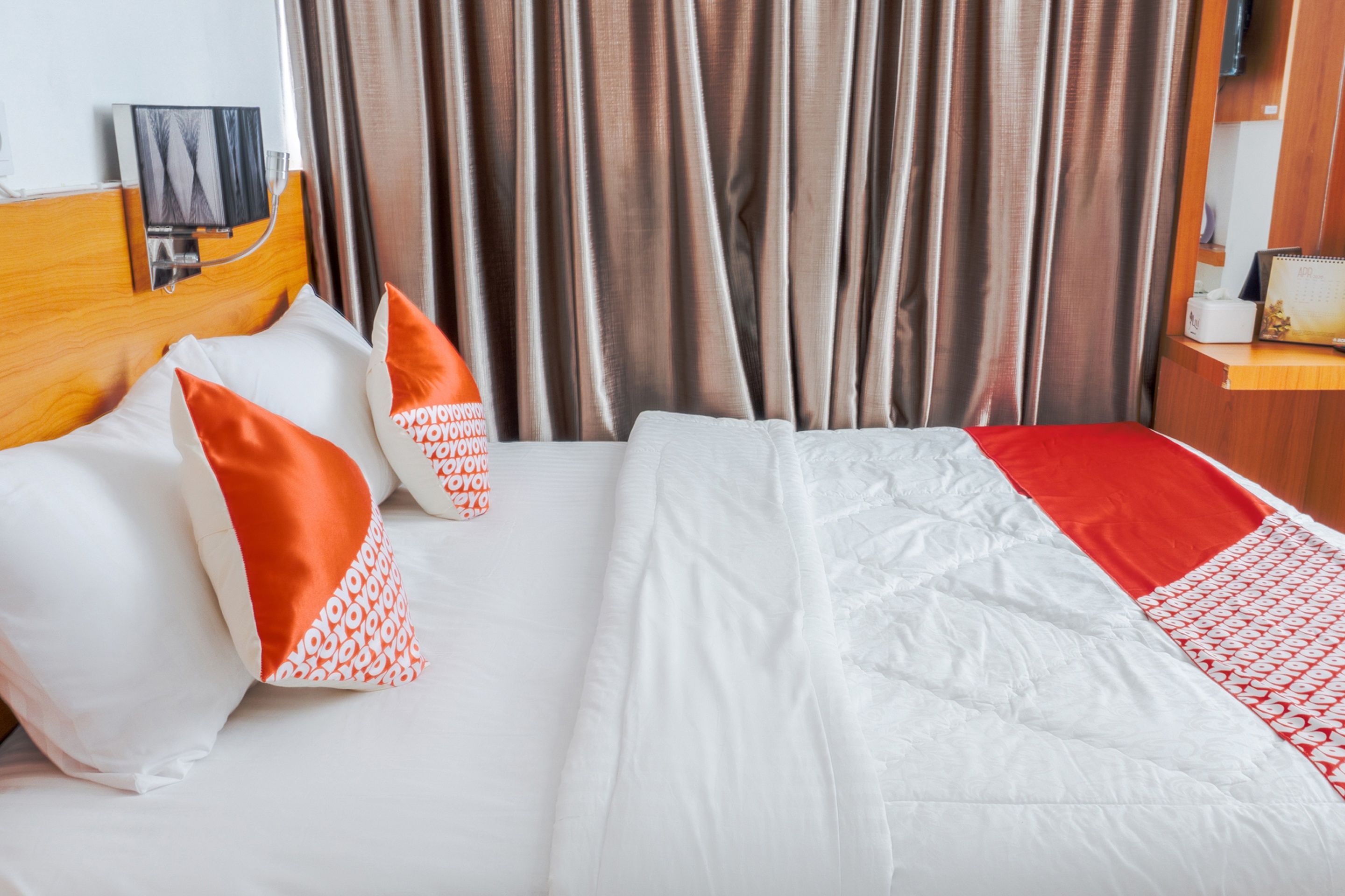 A hotel room with a bed, pillows, a headboard, curtains, and a bedside table with a lamp.