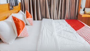 Habitación de hotel con cama, almohadas, cabecero, cortinas y mesita de noche con lámpara.