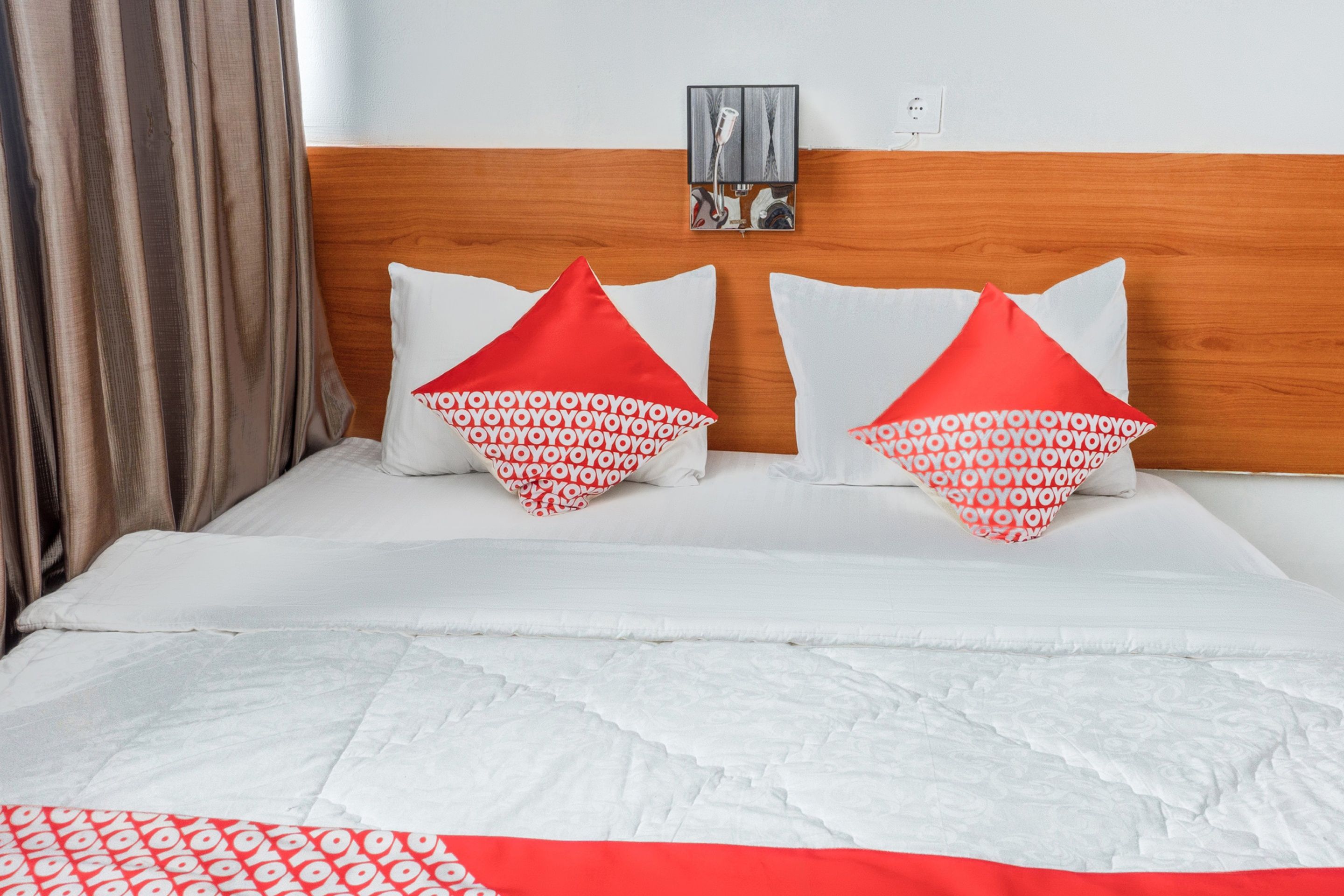 A hotel room with a wooden headboard, two pillows with a red and white pattern, a white quilt, and a red and white blanket.