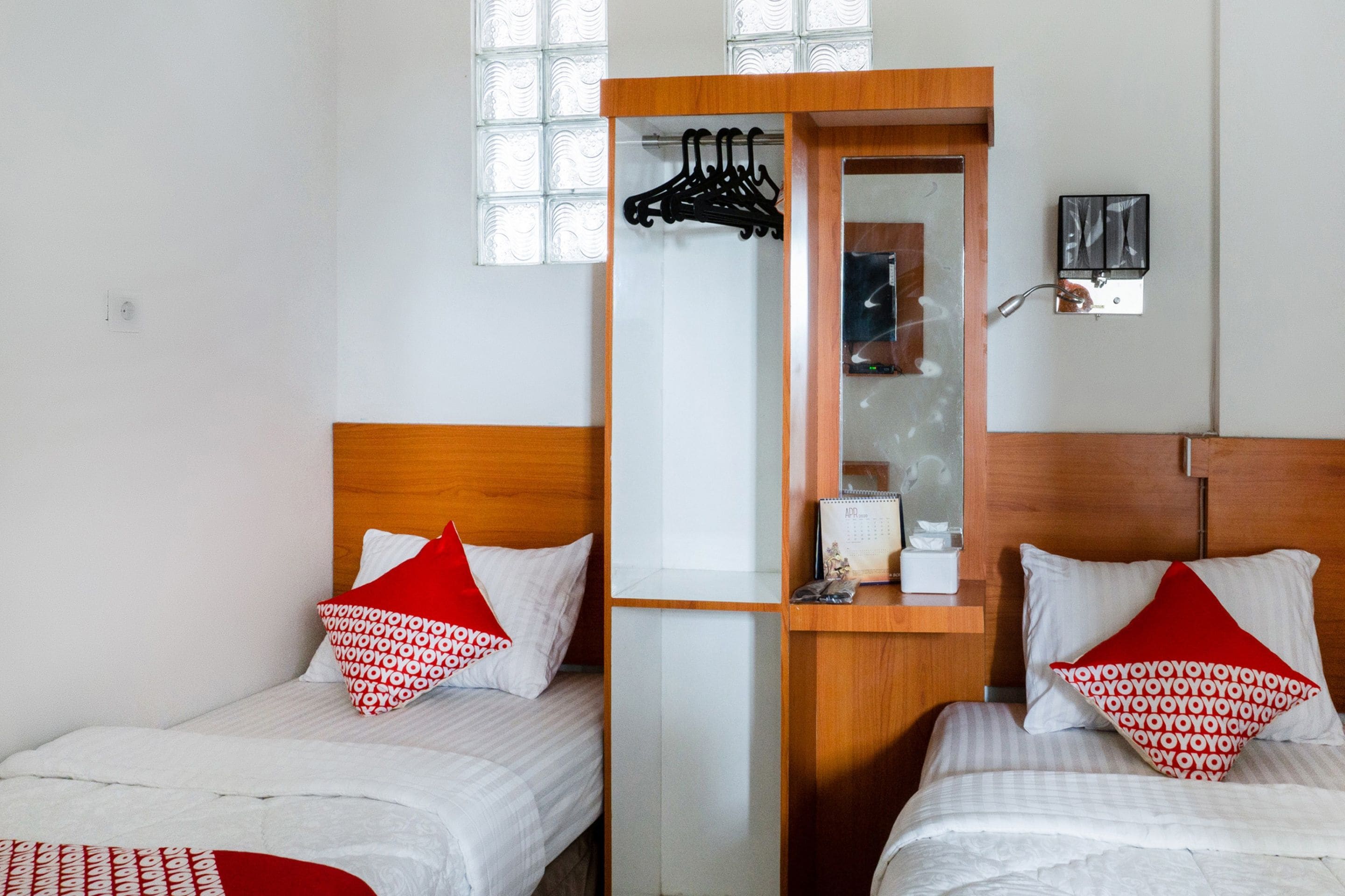 A hotel room with two beds, each with a red and white patterned pillow, a wooden headboard, and a small wooden desk with a mirror and a calendar.