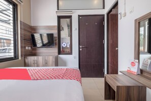 Quarto de hotel com cama, televisão na parede, guarda-roupa de madeira, porta escura e um banco.