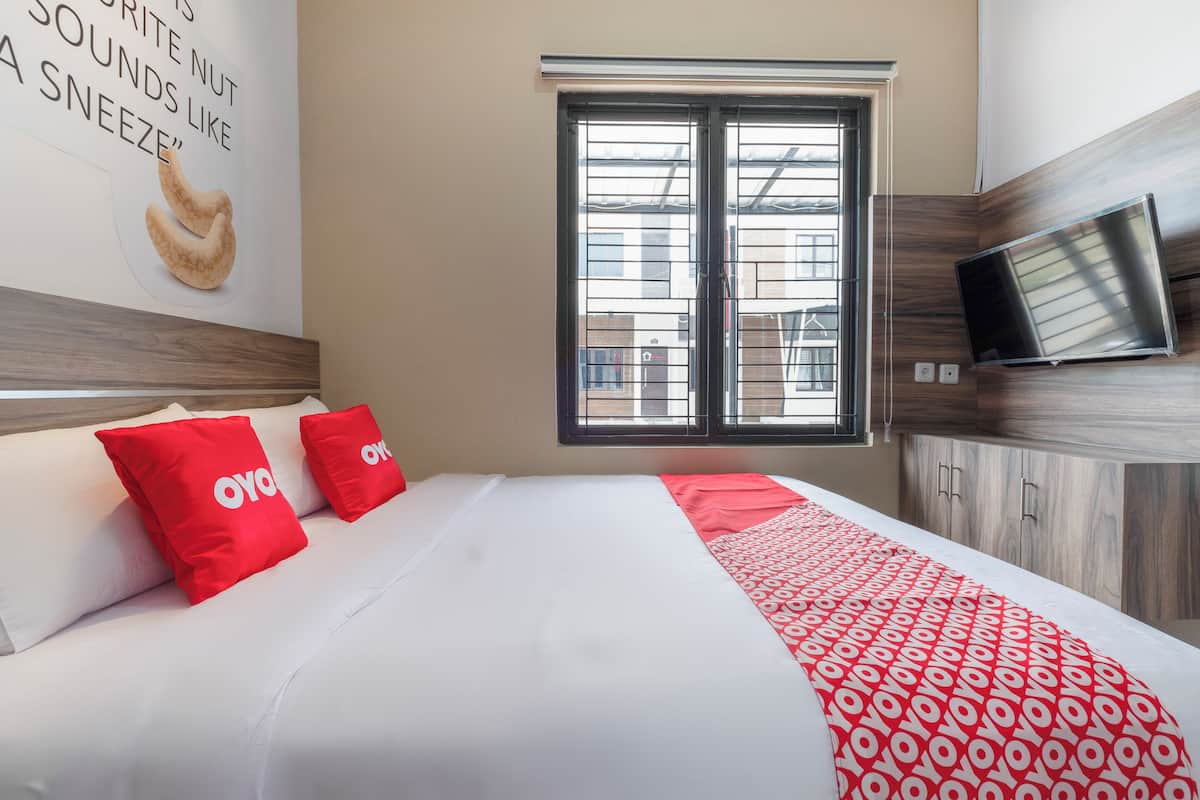 A hotel room with a bed, two red pillows, a patterned blanket, a television, and a window with a view of buildings.