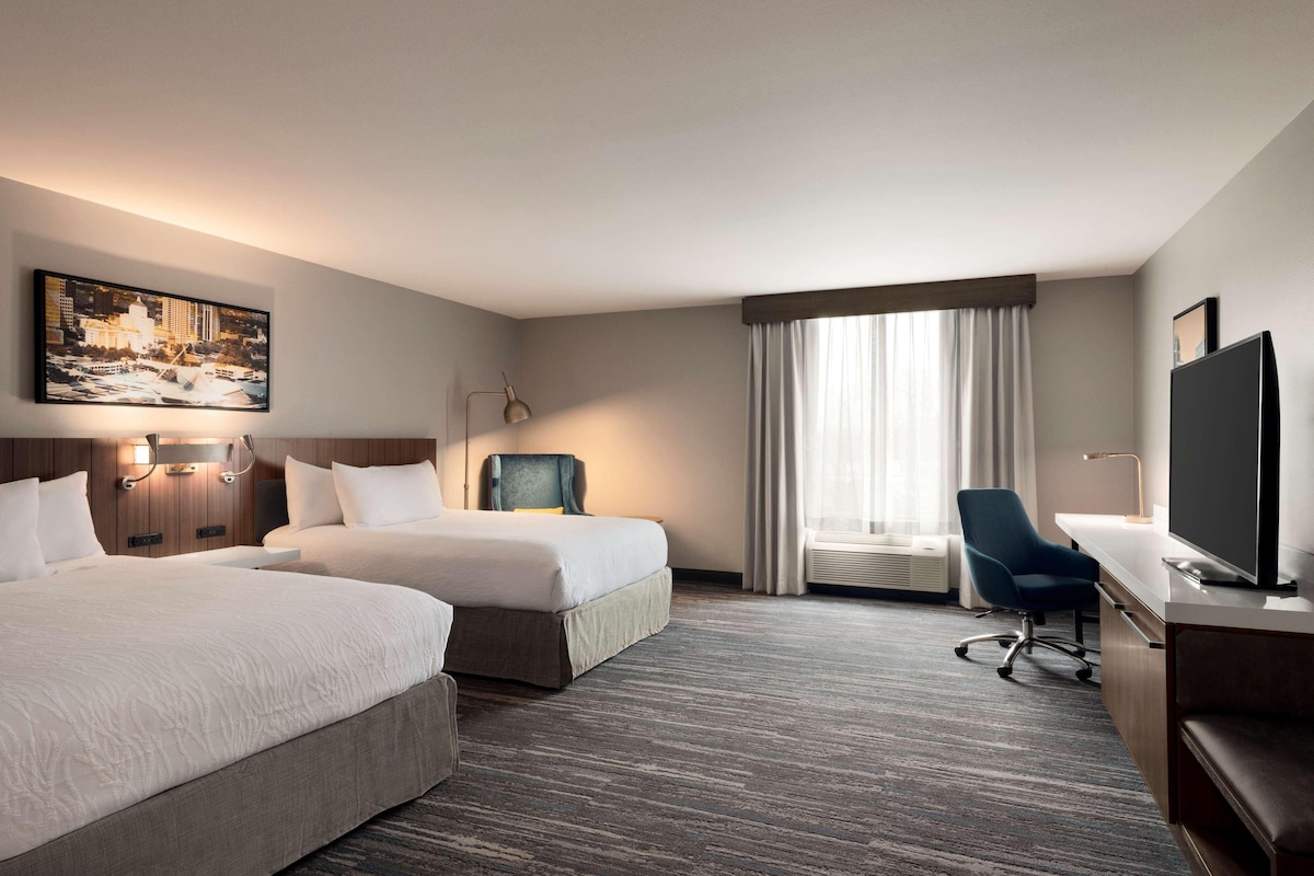 A hotel room with two beds, a desk, a chair, a television, and a large window with curtains.