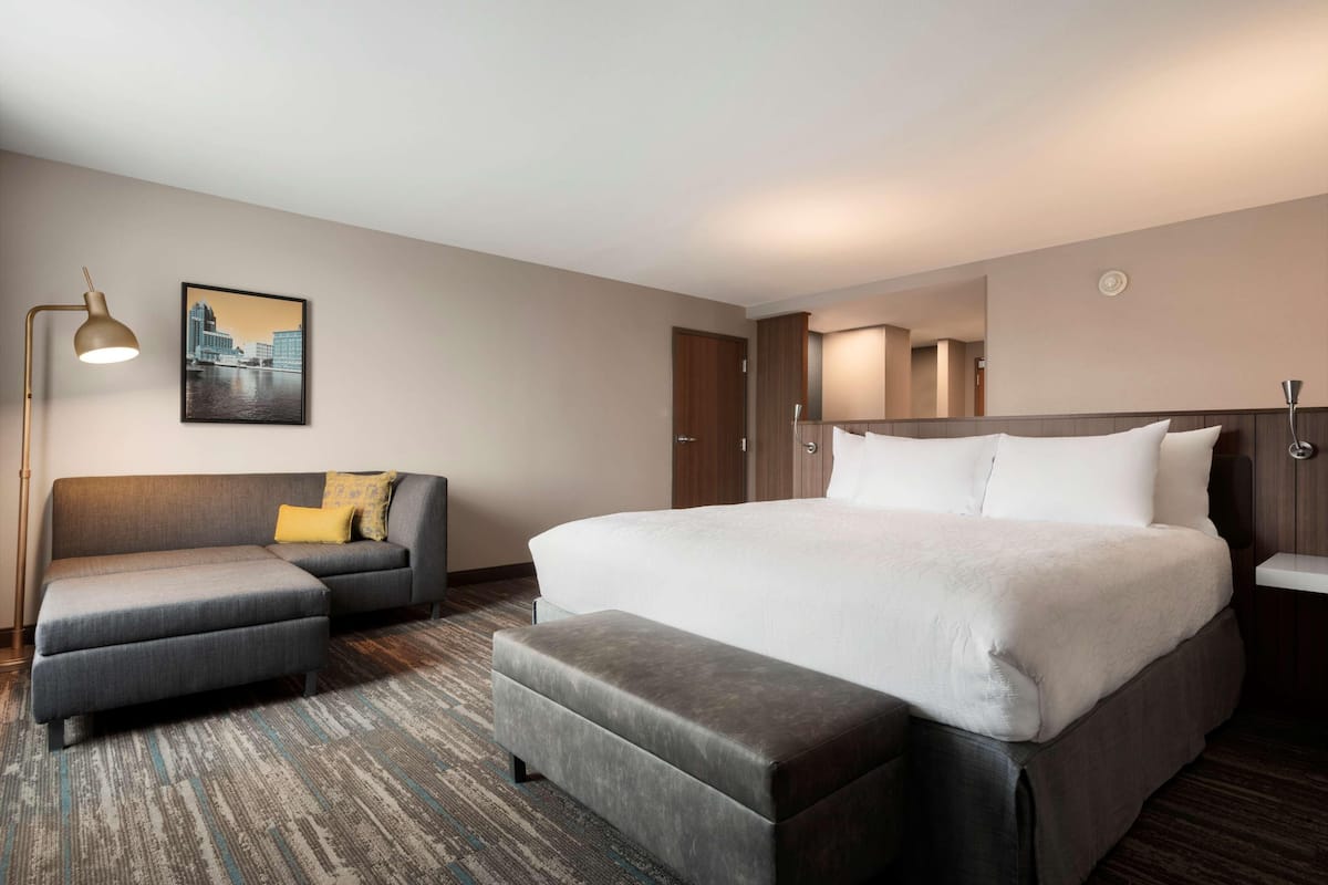 A modern hotel room with a large bed, a sofa, an ottoman, a desk, and a lamp.