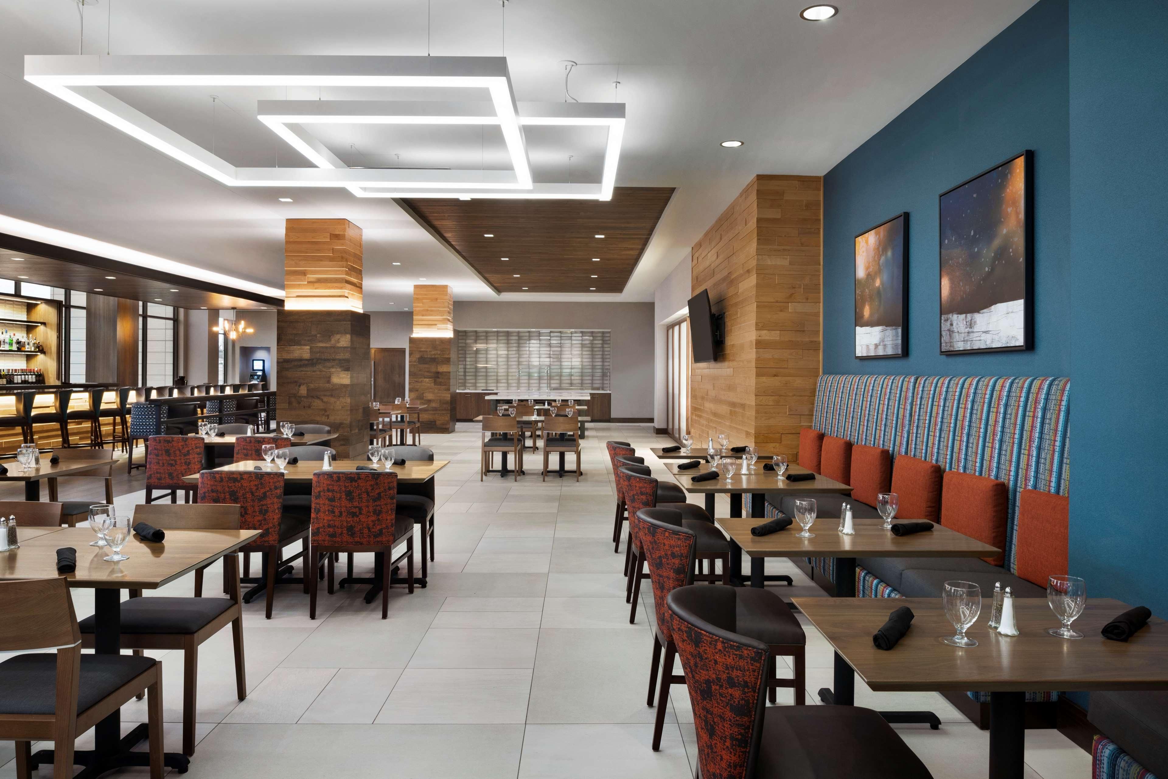 A modern restaurant with wooden tables and chairs, a bar area, and a seating section with cushioned benches.