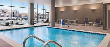 An indoor swimming pool with clear views of the outside, a seating area with tables and chairs, and a large window providing natural light.