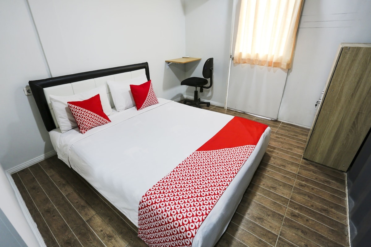 A neatly made bed with a white and red patterned bedspread, a black headboard, and two red pillows. There is a wooden shelf above the bed, a black office chair, and a wooden wardrobe.