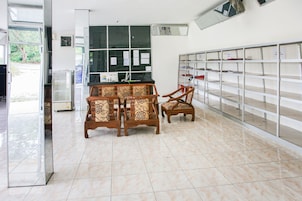 Um quarto com piso ladrilhado, uma mesa de madeira com cadeiras, uma janela com papéis e uma estante com compartimentos identificados.