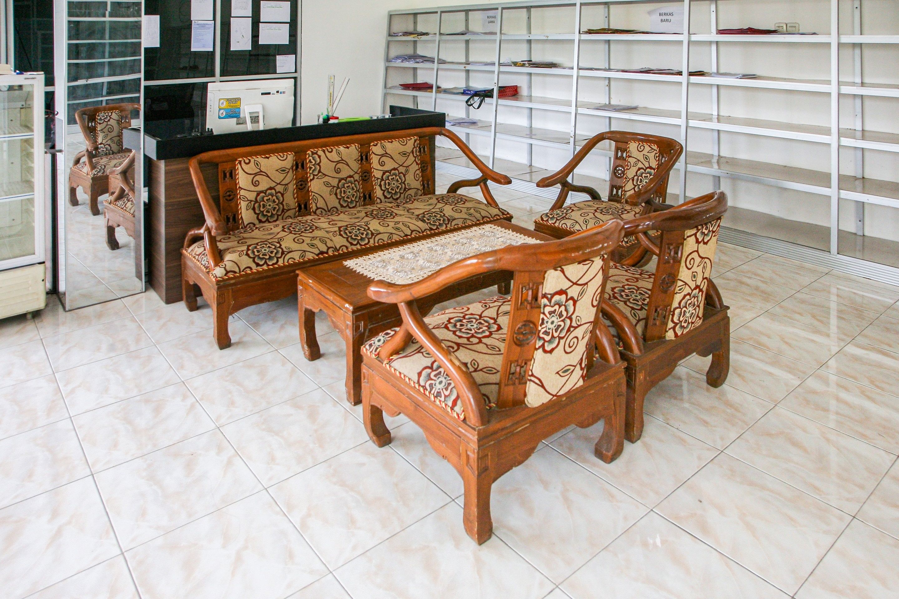 A room with a wooden sofa set, a small table, and a counter with a computer. Shelves with various items are in the background.