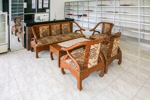 Quarto com sofá de madeira, uma mesa pequena e balcão com computador. Prateleiras com diversos objetos ao fundo.