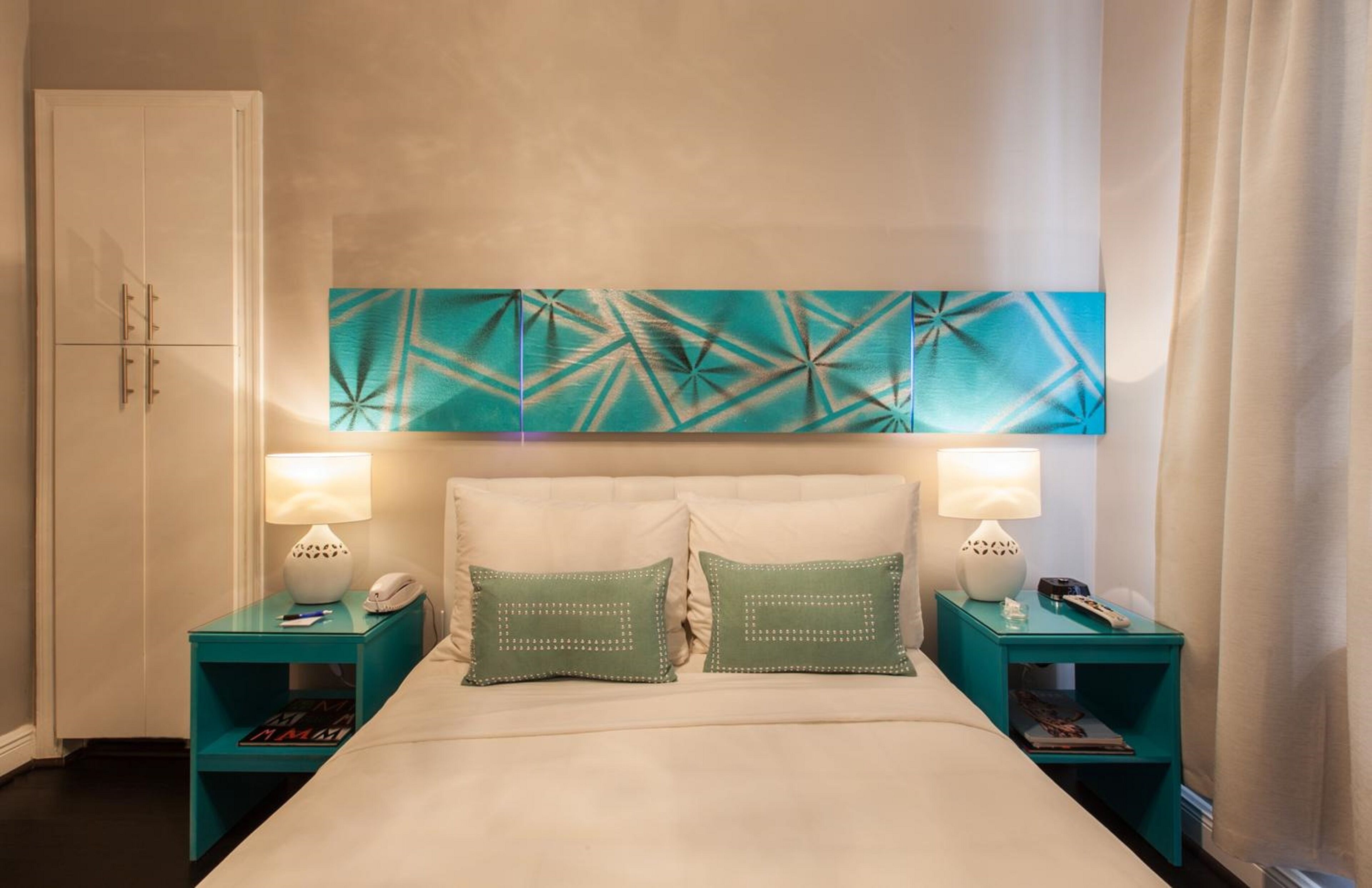 A hotel room with a bed, two nightstands, a white wardrobe, and a geometric wall art piece.
